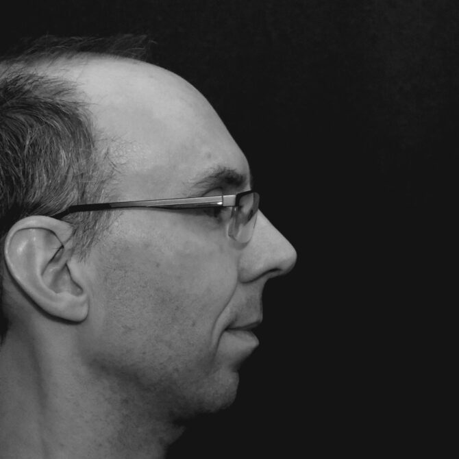 Schwarzweißfoto eines Mannes mittleren Alters mit Brille im Profil. Der Kopf schaut von links ins Bild, das rechte Drittel zeigt nur schwarzen Hintergrund.