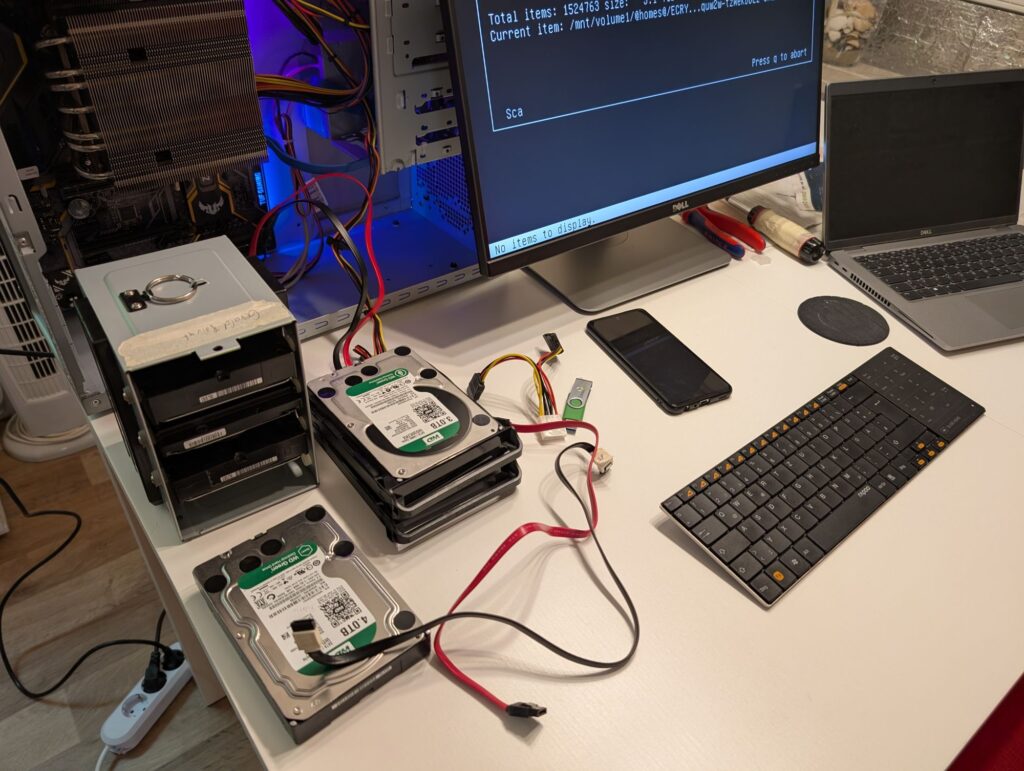 Auf einem Tisch steht ein Tower PC-Gehäuse mit offener Seitenwand. Davor steht ein Käfig mit drei Festplatten, daneben liegen drei weitere Lose Festplatten die teilweise am PC angeschlossen sind. Neben dem PC steht ein Monitor, Rechts davon noch ein aufgeklappter Laptop. Vor dem Monitor liegen ein Smartphone und eine Tastatur, darunter ein Schraubenzieher und eine Zange