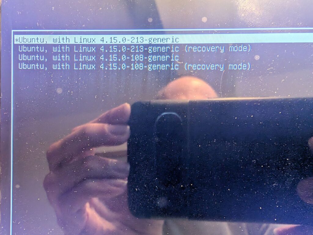 Ein Bildschirm wird abfotografiert, das Smartphone und der Mensch dahinter spiegeln sich im Bildschirm. Auf dem Bildschirm ist ein Grub Bootloader mit vier Einträgen zu sehen, "Ubuntu, with Linux 4.15.0.213-generic" und "Ubuntu, with Linux 4.15.0-108-generic" jeweils mit und ohne "recovery mode". Der erste Eintrag ist ausgewählt.