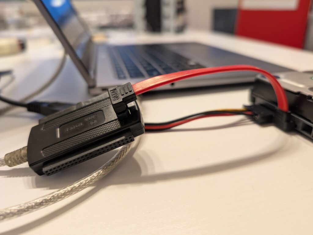 Neben einem aufgeklappten Laptop liegt eine Festplatte ohne Gehäuse. An die Festplatte mit einem kurzen roten SATA-Kabel schwarzer Adapter angeschlossen, der über ein silbernes USB-Kabel mit dem Laptop verbunden ist. Zum Stromanschluss der Festplatte geht ein Bund aus schwarz/rot/gelben Kabeln.